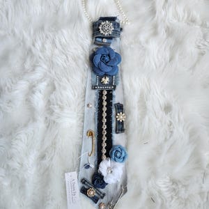 Puede incluir: Una corbata de mezclilla adornada con varios adornos, incluyendo una gran flor azul de tela, detalles de perlas y un broche decorativo. La corbata tambi&eacute;n presenta un imperdible y una etiqueta que dice "A Twist by Lee."