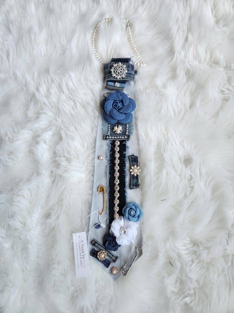 Puede incluir: Una corbata de mezclilla adornada con varios adornos, incluyendo una gran flor azul de tela, detalles de perlas y un broche decorativo. La corbata tambi&eacute;n presenta un imperdible y una etiqueta que dice "A Twist by Lee."