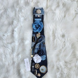 Puede incluir: Una corbata de mezclilla adornada con varios adornos, incluyendo flores de tela azul, perlas y alfileres decorativos. La corbata est&aacute; conectada a un collar de perlas. Una etiqueta con texto est&aacute; adjunta.