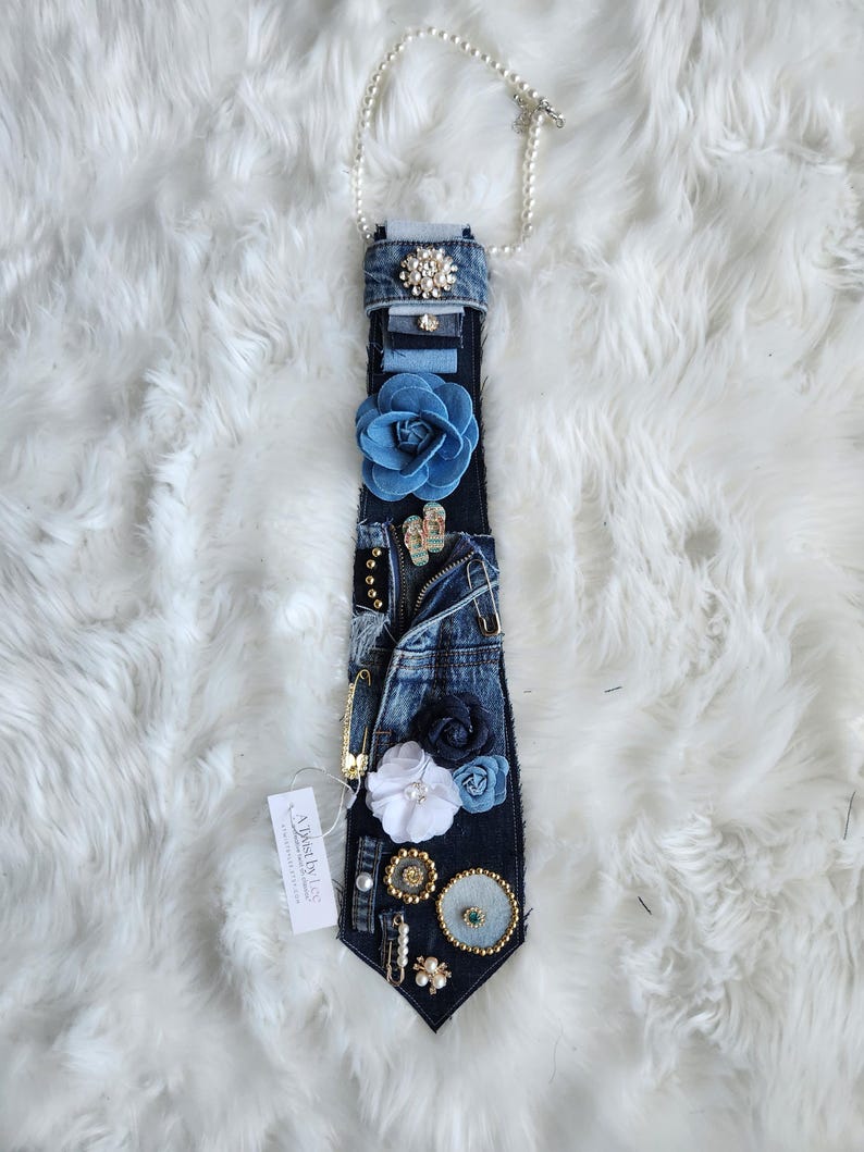 Puede incluir: Una corbata de mezclilla adornada con varios adornos, incluyendo flores de tela azul, perlas y alfileres decorativos. La corbata est&aacute; conectada a un collar de perlas. Una etiqueta con texto est&aacute; adjunta.