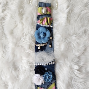 May include: A denim tie adorned with fabric flowers, pearls, and decorative embellishments. The tie features a blue flower, a green and pink striped accent, and a pearl necklace. The tie is on a white fluffy surface.