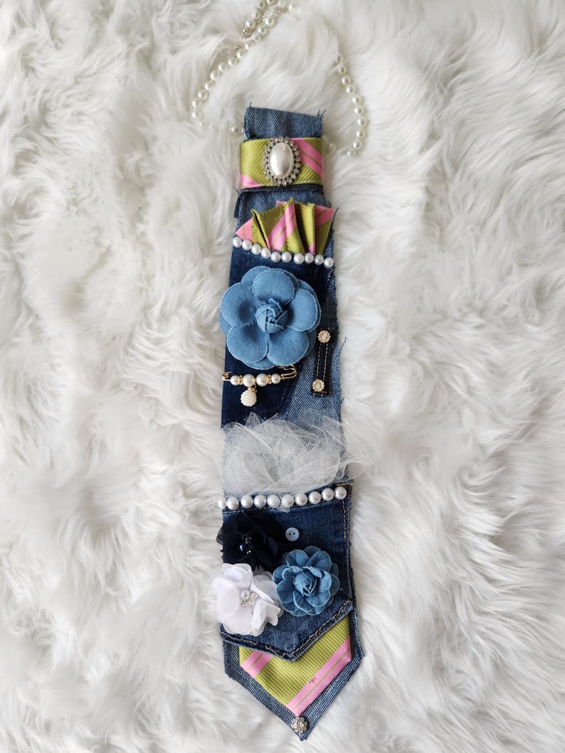 May include: A denim tie adorned with fabric flowers, pearls, and decorative embellishments. The tie features a blue flower, a green and pink striped accent, and a pearl necklace. The tie is on a white fluffy surface.