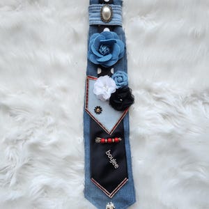 May include: A denim tie adorned with fabric flowers in blue, white, and black. The tie features a black section with the word "boujee" and is embellished with a pearl and rhinestone accents. The tie is set against a white, fluffy background.