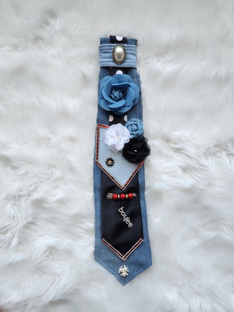 May include: A denim tie adorned with fabric flowers in blue, white, and black. The tie features a black section with the word "boujee" and is embellished with a pearl and rhinestone accents. The tie is set against a white, fluffy background.