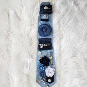 Puede incluir: Un collar de corbata de mezclilla con varios adornos. La corbata azul claro presenta bolsillos de mezclilla azul oscuro, una gran flor de tela azul y varias flores de tela m&aacute;s peque&ntilde;as en blanco y azul. Est&aacute; adornada con perlas, pedrer&iacute;a y una cadena plateada.