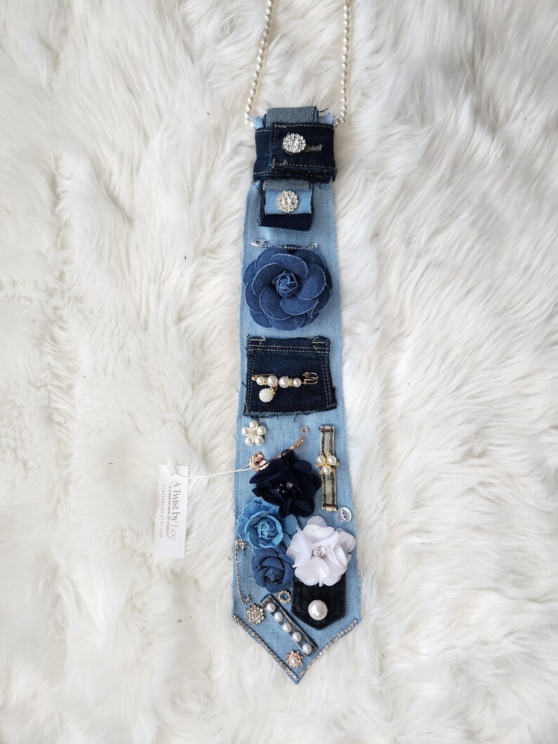 Puede incluir: Un collar de corbata de mezclilla con varios adornos. La corbata azul claro presenta bolsillos de mezclilla azul oscuro, una gran flor de tela azul y varias flores de tela m&aacute;s peque&ntilde;as en blanco y azul. Est&aacute; adornada con perlas, pedrer&iacute;a y una cadena plateada.