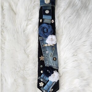 Puede incluir: Una corbata de mezclilla hecha a mano con varios parches de mezclilla, adornada con flores de tela, perlas y alfileres decorativos. La corbata presenta una base de mezclilla oscura con detalles de mezclilla m&aacute;s claros y una variedad de elementos decorativos.