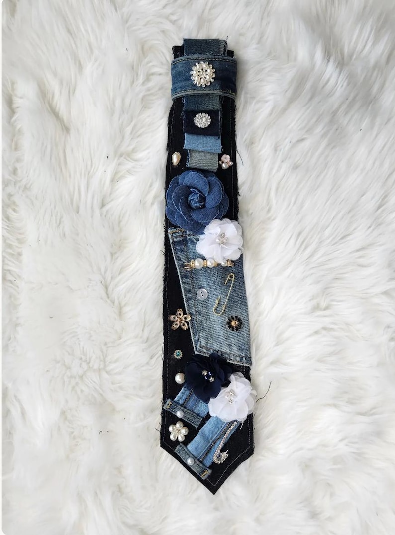 Puede incluir: Una corbata de mezclilla hecha a mano con varios parches de mezclilla, adornada con flores de tela, perlas y alfileres decorativos. La corbata presenta una base de mezclilla oscura con detalles de mezclilla m&aacute;s claros y una variedad de elementos decorativos.