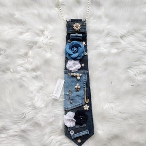 Puede incluir: Una corbata de mezclilla adornada con varios adornos, que incluyen flores de tela azules y blancas, perlas y broches brillantes. Una cadena de perlas est&aacute; unida en la parte superior. La etiqueta dice "A Twist by Liz".