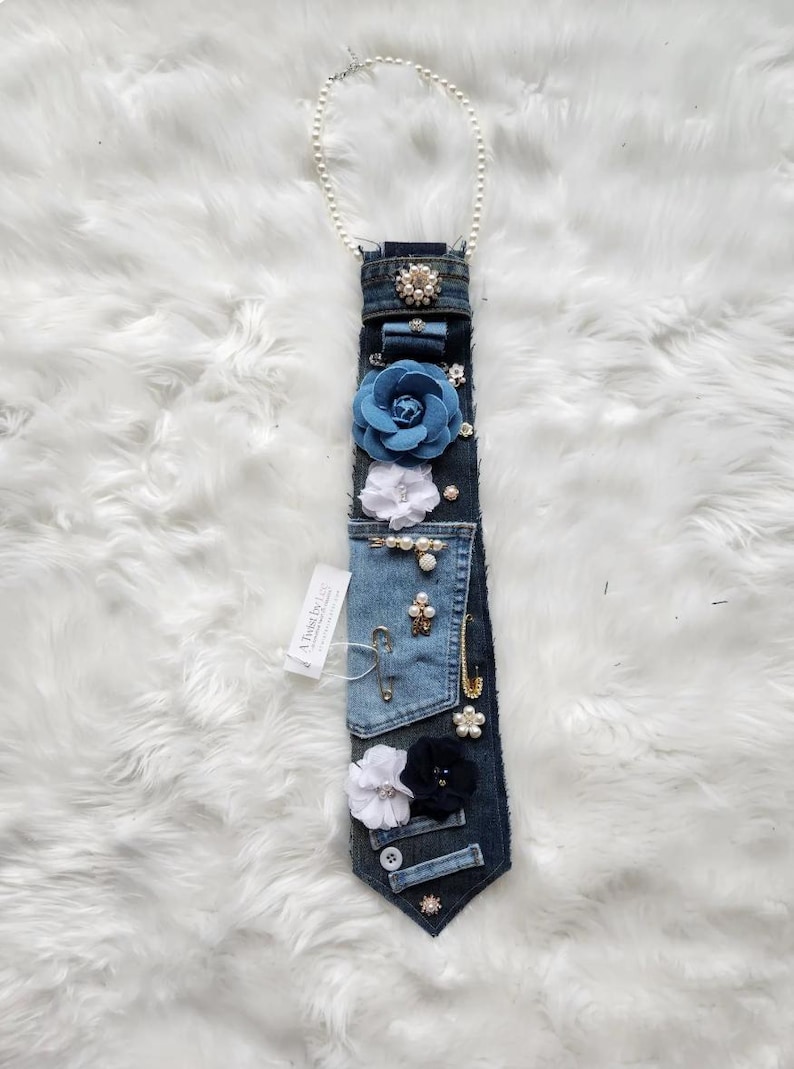 Puede incluir: Una corbata de mezclilla adornada con varios adornos, que incluyen flores de tela azules y blancas, perlas y broches brillantes. Una cadena de perlas est&aacute; unida en la parte superior. La etiqueta dice "A Twist by Liz".