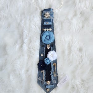 Puede incluir: Corbata de mezclilla adornada con varios elementos decorativos. Incluye un collar de perlas, detalles florales azules y blancos, y broches brillantes. La corbata est&aacute; adornada con imperdibles y otros detalles.