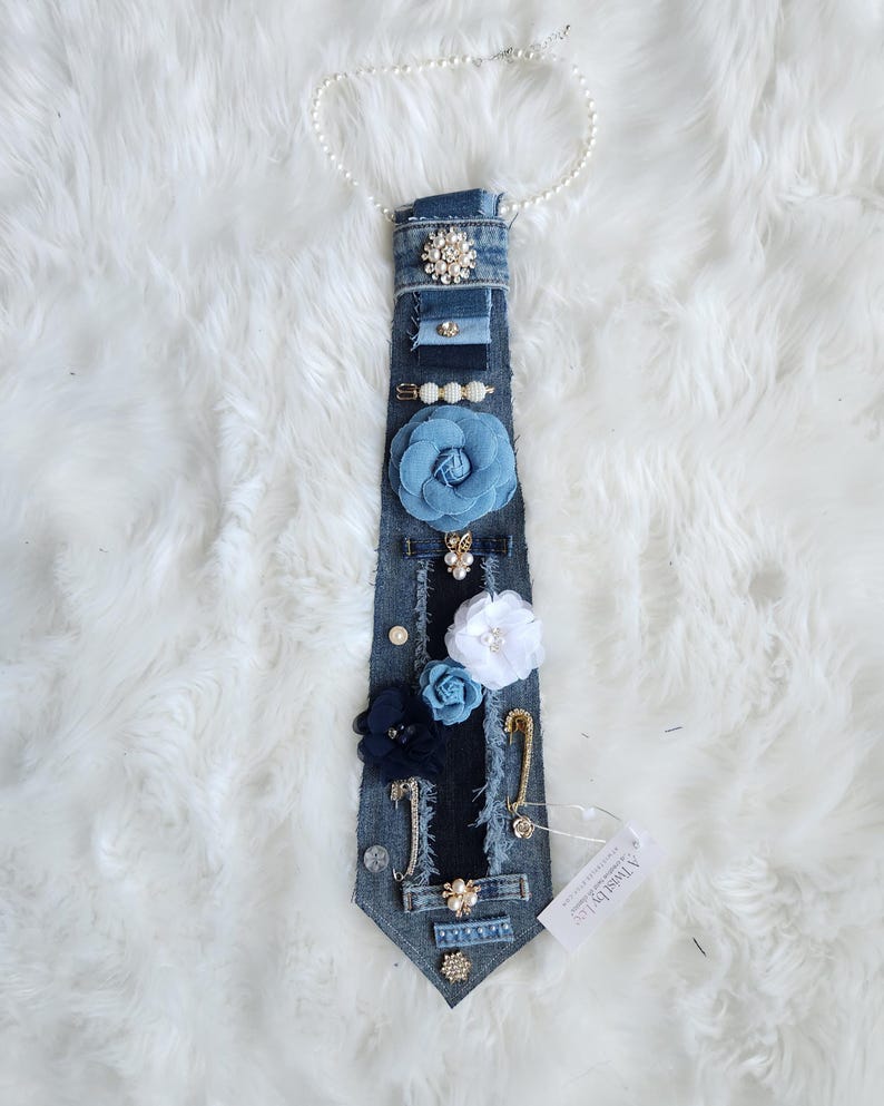 Puede incluir: Corbata de mezclilla adornada con varios elementos decorativos. Incluye un collar de perlas, detalles florales azules y blancos, y broches brillantes. La corbata est&aacute; adornada con imperdibles y otros detalles.