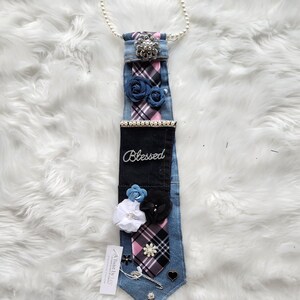 May include: A denim tie adorned with fabric flowers, a pearl necklace, and a decorative brooch. The tie features a plaid pattern in pink, black, and white, with the word "Blessed" embroidered on it. Additional embellishments include a heart-shaped charm and a decorative pin.