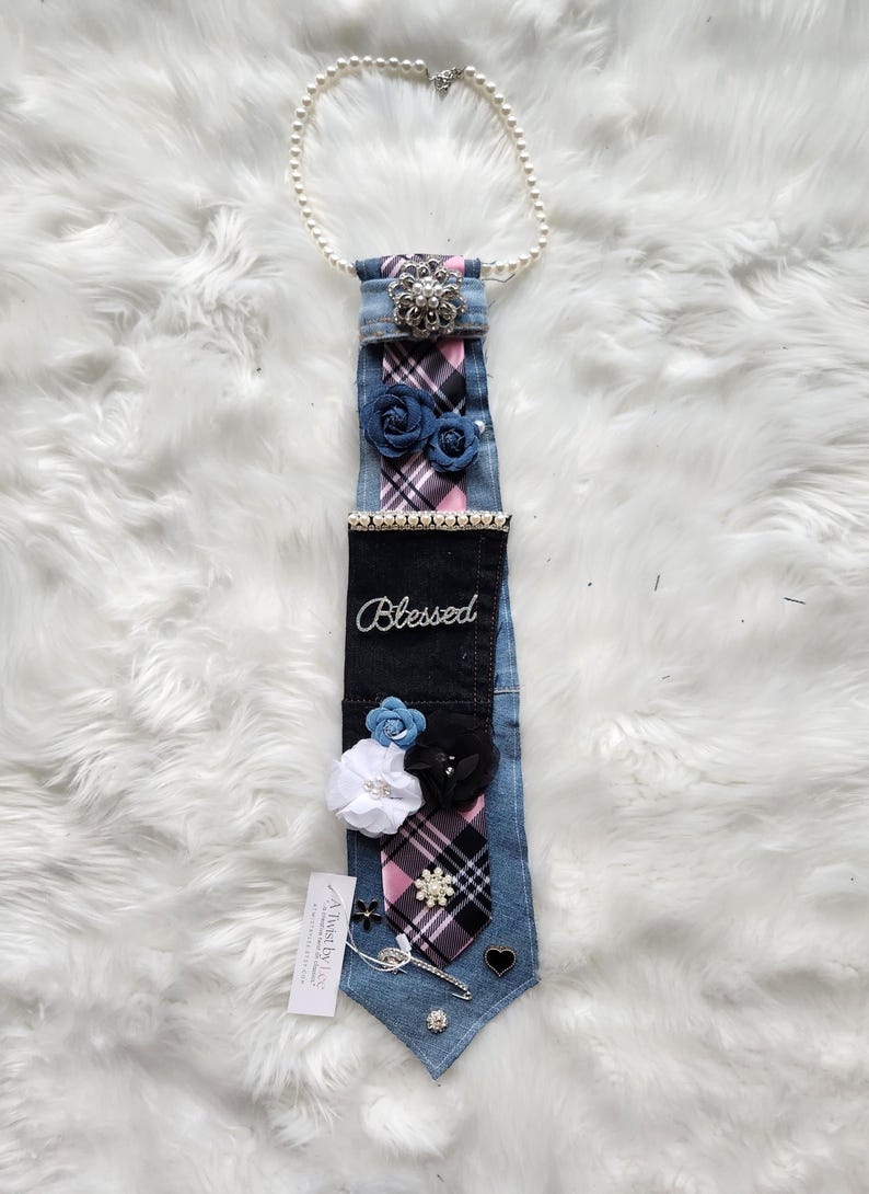 May include: A denim tie adorned with fabric flowers, a pearl necklace, and a decorative brooch. The tie features a plaid pattern in pink, black, and white, with the word "Blessed" embroidered on it. Additional embellishments include a heart-shaped charm and a decorative pin.