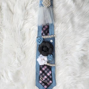 May include: A denim tie adorned with a pearl and rhinestone embellishment, a sheer tulle accent, and fabric flowers in black, white, and blue. The tie features a plaid pattern and a decorative safety pin.