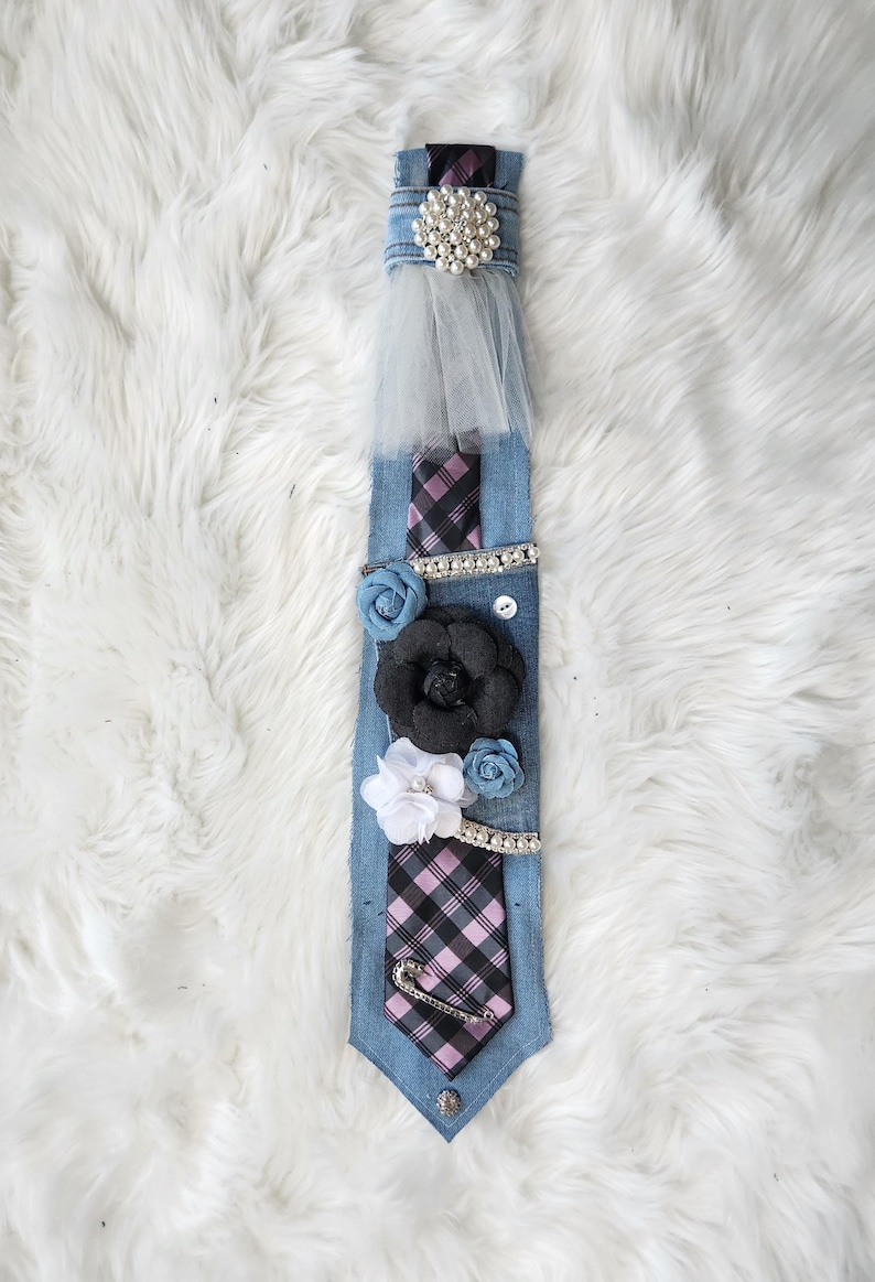May include: A denim tie adorned with a pearl and rhinestone embellishment, a sheer tulle accent, and fabric flowers in black, white, and blue. The tie features a plaid pattern and a decorative safety pin.