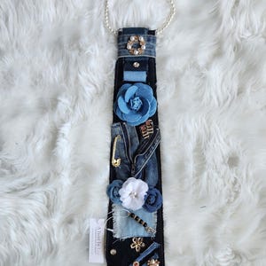 Puede incluir: Corbata de mezclilla adornada con flores de tela azul, un imperdible dorado y detalles de perlas. La corbata est&aacute; adornada con un collar de perlas y una etiqueta que dice "A Twist by Lee".