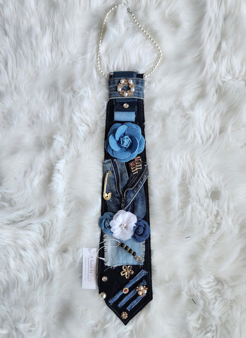 Puede incluir: Corbata de mezclilla adornada con flores de tela azul, un imperdible dorado y detalles de perlas. La corbata est&aacute; adornada con un collar de perlas y una etiqueta que dice "A Twist by Lee".