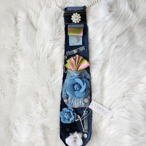 May include: A denim tie adorned with fabric flowers, ribbons, and pearl accents. The tie features a variety of embellishments, including a large blue flower, a pearl necklace, and a tag that reads "A Twist by Lee."