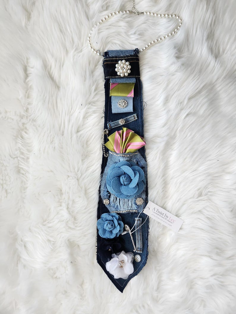 May include: A denim tie adorned with fabric flowers, ribbons, and pearl accents. The tie features a variety of embellishments, including a large blue flower, a pearl necklace, and a tag that reads "A Twist by Lee."