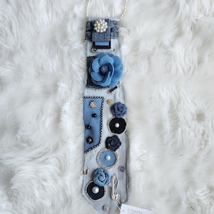 Puede incluir: Una corbata de mezclilla adornada con varios adornos, que incluyen un collar de perlas, una gran flor de tela azul y flores decorativas m&aacute;s peque&ntilde;as. La corbata presenta una variedad de elementos decorativos, incluyendo perlas, pedrer&iacute;a y imperdibles.