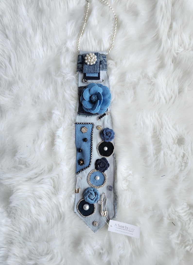 Puede incluir: Una corbata de mezclilla adornada con varios adornos, que incluyen un collar de perlas, una gran flor de tela azul y flores decorativas m&aacute;s peque&ntilde;as. La corbata presenta una variedad de elementos decorativos, incluyendo perlas, pedrer&iacute;a y imperdibles.