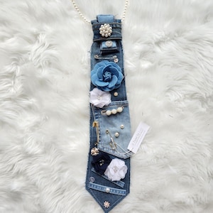 Puede incluir: Una corbata de mezclilla adornada con varios adornos. Presenta una gran flor de tela azul, flores de tela blanca y azul marino, perlas y alfileres decorativos. Una cadena de perlas est&aacute; unida en la parte superior. La etiqueta dice "A Twist by Lee".