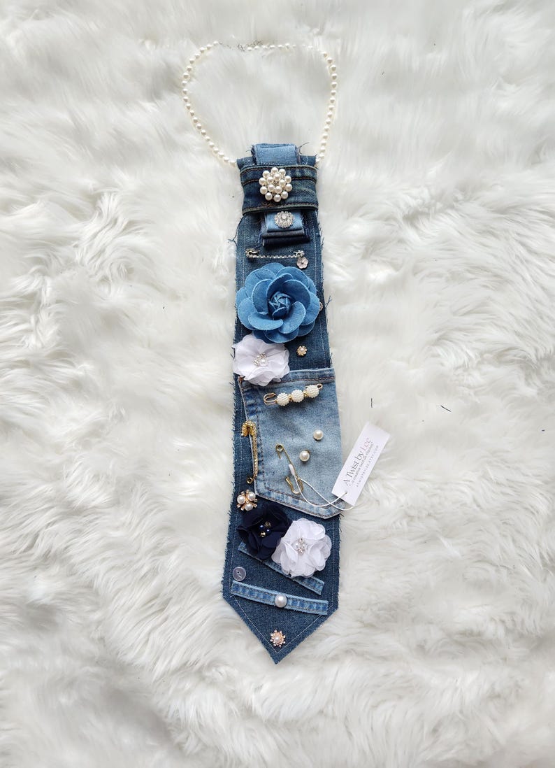 Puede incluir: Una corbata de mezclilla adornada con varios adornos. Presenta una gran flor de tela azul, flores de tela blanca y azul marino, perlas y alfileres decorativos. Una cadena de perlas est&aacute; unida en la parte superior. La etiqueta dice "A Twist by Lee".