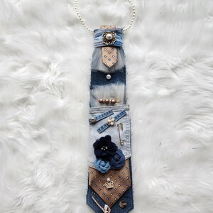 May include: A unique, handmade denim tie adorned with various embellishments. Features a pearl necklace, fabric flowers, decorative pins, and a small tag that reads "A Twist by Lee". The tie has a brown and blue patterned design.