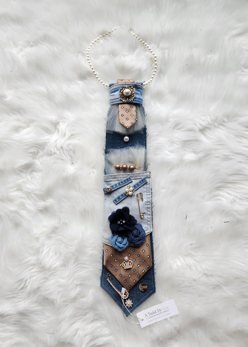 May include: A unique, handmade denim tie adorned with various embellishments. Features a pearl necklace, fabric flowers, decorative pins, and a small tag that reads "A Twist by Lee". The tie has a brown and blue patterned design.