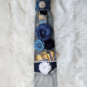 May include: A denim tie adorned with various embellishments. It features a pearl necklace, a large rhinestone brooch, and several fabric flowers in shades of blue and white. Gold and white striped ribbons and a white tulle accent add texture.
