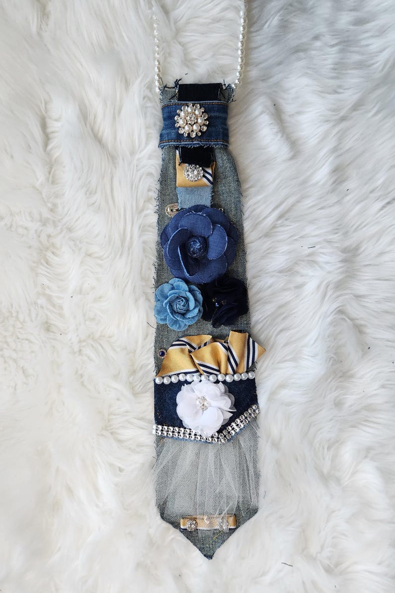 May include: A denim tie adorned with various embellishments. It features a pearl necklace, a large rhinestone brooch, and several fabric flowers in shades of blue and white. Gold and white striped ribbons and a white tulle accent add texture.