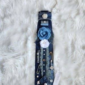 Puede incluir: Una corbata de mezclilla adornada con varios adornos, incluyendo detalles de perlas, flores de tela y alfileres decorativos. La corbata presenta un collar de perlas y una etiqueta que dice "A Twist by Liz".