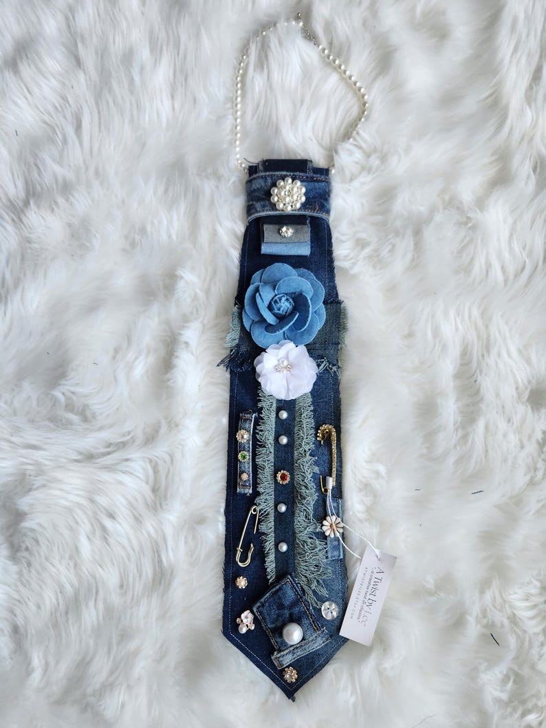 Puede incluir: Una corbata de mezclilla adornada con varios adornos, incluyendo detalles de perlas, flores de tela y alfileres decorativos. La corbata presenta un collar de perlas y una etiqueta que dice "A Twist by Liz".