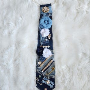Puede incluir: Corbata de mezclilla adornada con flores de tela, perlas y alfileres decorativos. La corbata presenta un collar de perlas en la parte superior. El dise&ntilde;o incorpora detalles en azul, blanco y dorado. La etiqueta dice "A Twist by Les".