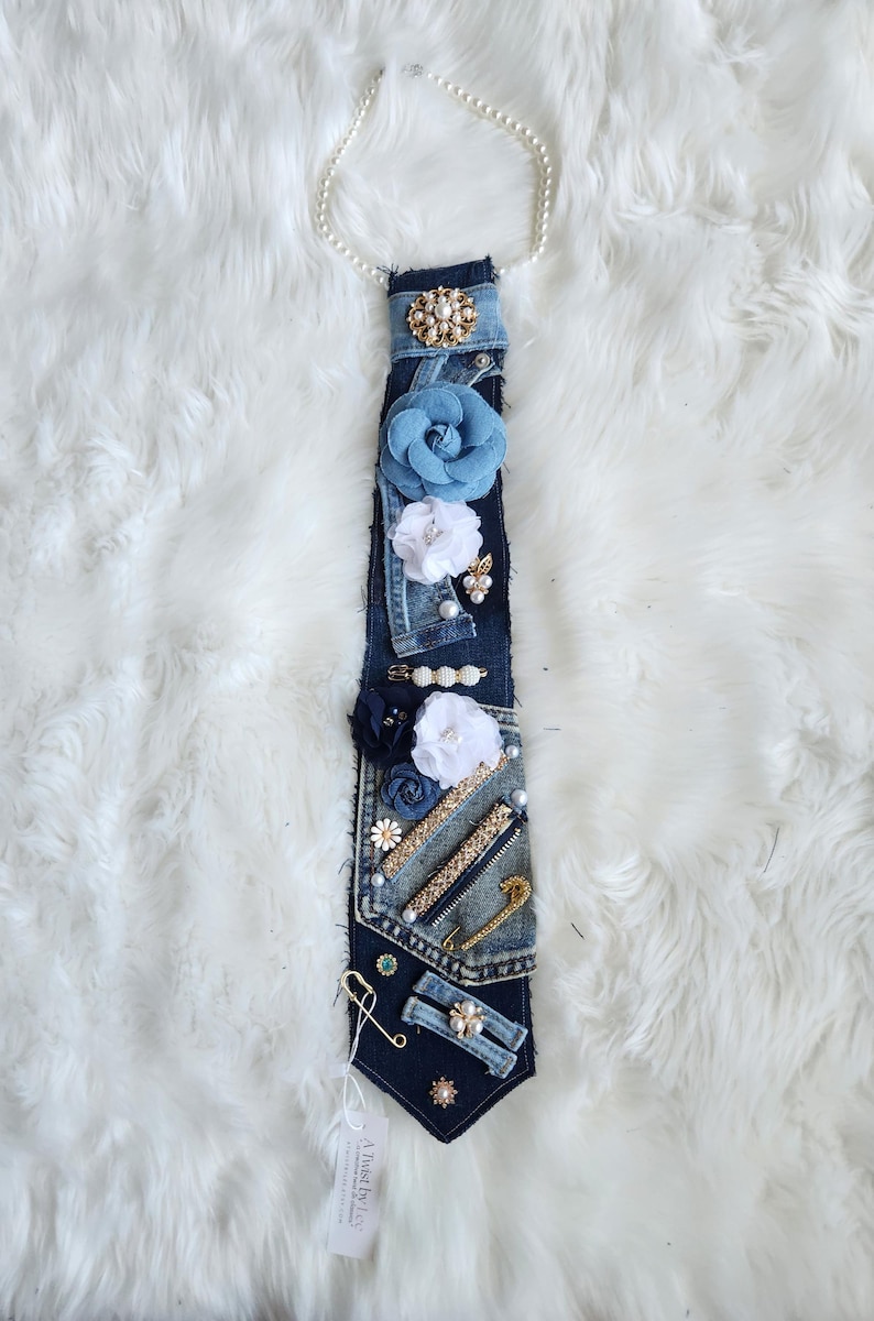 Puede incluir: Corbata de mezclilla adornada con flores de tela, perlas y alfileres decorativos. La corbata presenta un collar de perlas en la parte superior. El dise&ntilde;o incorpora detalles en azul, blanco y dorado. La etiqueta dice "A Twist by Les".