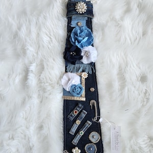 Puede incluir: Una corbata de mezclilla adornada con flores de tela en tonos azules y blancos, adornada con pedrer&iacute;a, perlas y alfileres decorativos. La corbata est&aacute; unida a un collar de perlas.