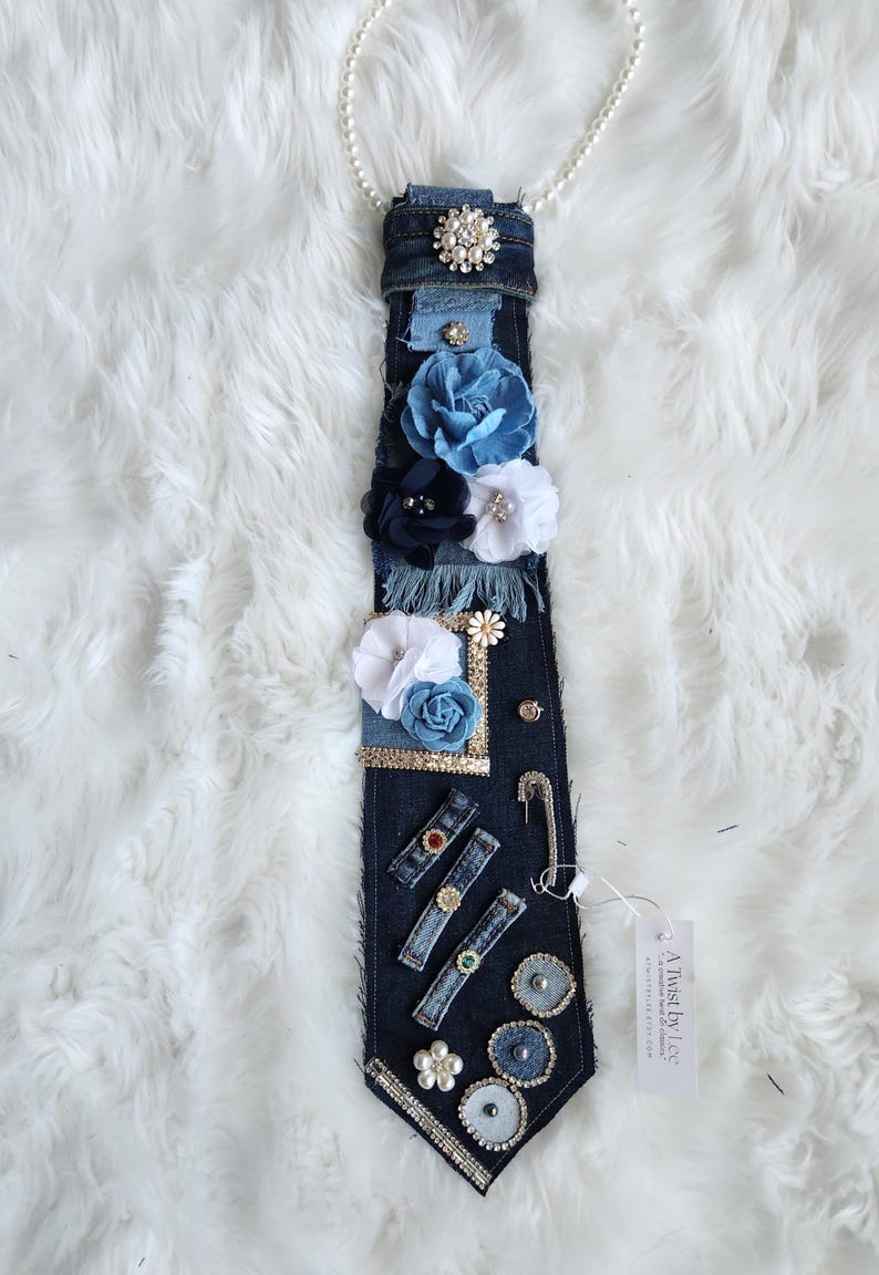Puede incluir: Una corbata de mezclilla adornada con flores de tela en tonos azules y blancos, adornada con pedrer&iacute;a, perlas y alfileres decorativos. La corbata est&aacute; unida a un collar de perlas.