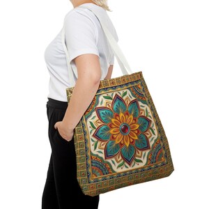 May include: A tote bag featuring a large, symmetrical mandala design in shades of teal, yellow, and deep red. The pattern is framed by a gold-toned border with a grid of small squares and red accents, set against a cream-colored background.