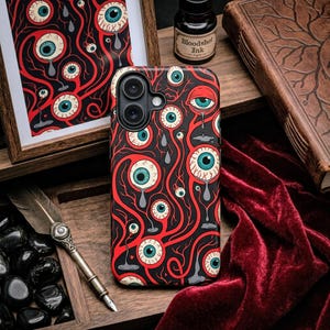 May include: A black phone case with a repeating pattern of red veins, teal eyes, and teardrops. A framed print with the same design is visible. A bottle of "Bloodshot Ink" and a vintage book are also in the image.