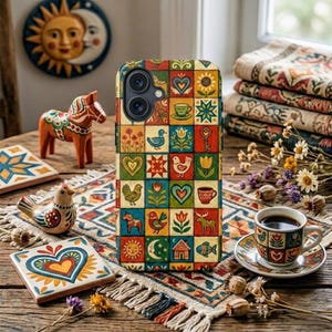 May include: A patterned phone case with a patchwork design featuring hearts, birds, flowers, and other folk art motifs. The case is on a wooden table with a matching coffee cup and saucer, and other decorative items.