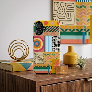 May include: A yellow smartphone case with a colorful geometric pattern. The case features a patchwork design with stripes, circles, and abstract shapes in various colors. A gold-colored decorative object and a small yellow vase are also visible.