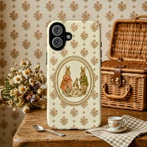 May include: A cream-colored phone case featuring a vintage illustration of a rabbit family in an oval frame. The case has a floral pattern and is set on a wooden table with a picnic basket and teacup in the background.