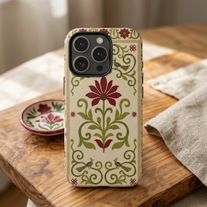 May include: A beige phone case with a floral design featuring a large burgundy flower, green leaves, and decorative birds. The case is on a wooden surface next to a small matching dish with a similar floral pattern.
