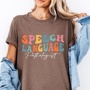 May include: Heather brown t-shirt with the words "SPEECH LANGUAGE Pathologist" in colorful, retro-style lettering. The shirt has rolled-up sleeves and a relaxed fit.