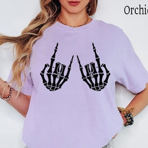 May include: A light purple short-sleeve t-shirt featuring a black graphic print of two skeleton hands making the rock and roll hand gesture. The text "Orchid" is printed in the upper right corner of the garment, which has a crew neckline.