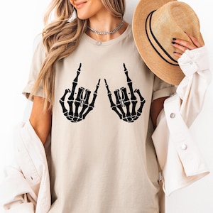 May include: A sand-colored t-shirt featuring a black graphic of two skeleton hands making the rock and roll sign. The shirt is paired with a light-colored button-down shirt and a tan fedora hat with a black band. The text Sand is visible.