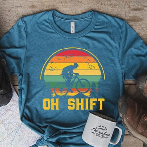 May include: Teal t-shirt with a colorful sunset design featuring a cyclist and the words "OH SHIFT." A white mug with the text "And so the adventure begins" sits next to the shirt. Brown boots and a backpack are also visible.