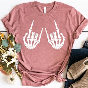 May include: A dusty rose short-sleeve t-shirt featuring a white graphic print of two skeletal hands making the rock and roll sign. The shirt has a crew neckline and is displayed flat with a bouquet of dried flowers, blue jeans, and beige sandals.
