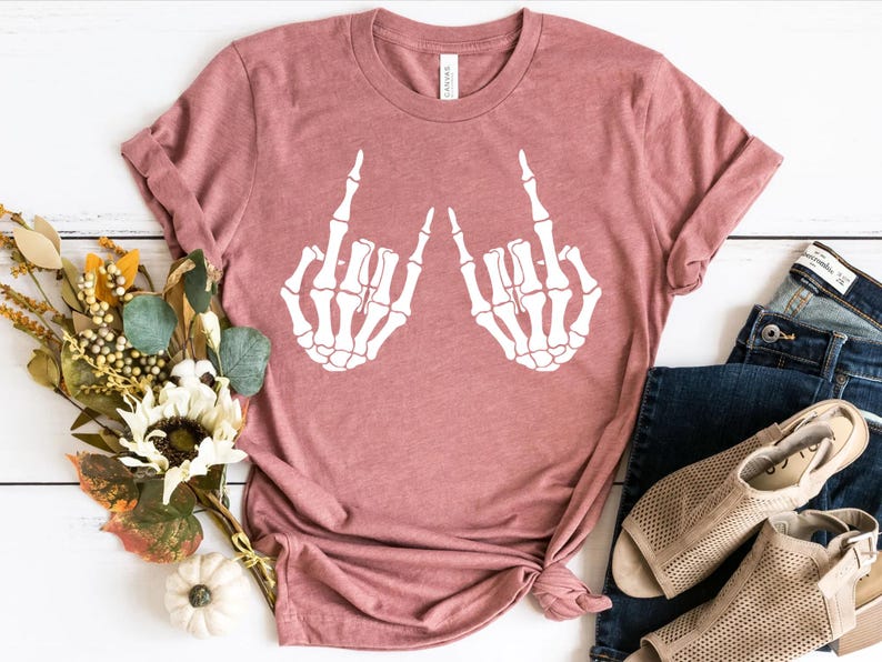 May include: A dusty rose short-sleeve t-shirt featuring a white graphic print of two skeletal hands making the rock and roll sign. The shirt has a crew neckline and is displayed flat with a bouquet of dried flowers, blue jeans, and beige sandals.